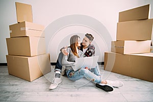 Young couple in love sitting on the floor of their new apartment, planning redecoration and searching for ideas on a laptop comput