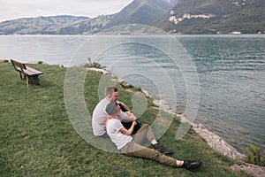young couple in love sitting on grass and looking at majestic mountain lake