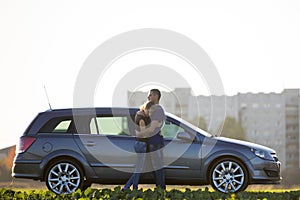 Young couple in love, slim attractive woman with long ponytail and handsome man standing together at silver car in green field on