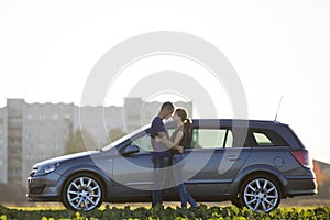 Young couple in love, slim attractive woman with long ponytail and handsome man standing together at silver car in green field on