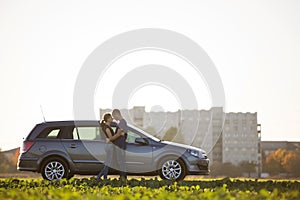 Young couple in love, slim attractive woman with long ponytail and handsome man standing together at silver car in green field on