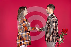 Young couple in love in the studio on a red background hold hands. The man hides the roses behind his back