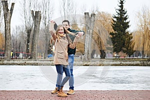 Young couple in love, wearing beige and green casual jackets, standing in front of frozen lake holding each others` hands, dancin