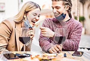 Young couple in love wearing open face masks having fun at wine bar outdoor - Happy traveler friends enjoying lunch together