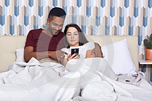 Young couple lying in bed, female iholding mobile phone, husband hugs his wife, watching film online, having rest together,