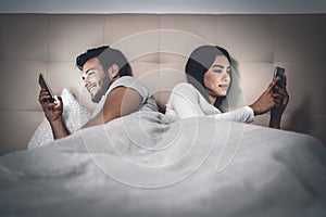 Young couple lying in bed using mobile phone while ignoring each other - Man and woman addicted to smartphone social networks