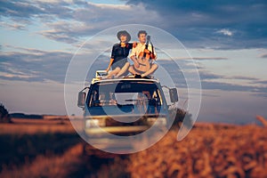 Young couple man with a guitar and woman in a hat are sitting on the roof of a car in a wheat field. Travel and adventur