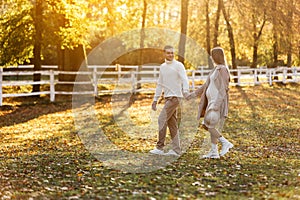 Young couple, man and pregnant wife are holding hands and walking in the autumn park on sunset. Happy family