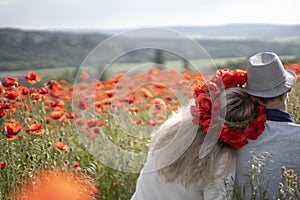 A young couple of man and woman in bright clothes are sitting with their backs to the audience in an embrace on a flowering poppy