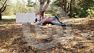 Young couple, man and woman having picnic in forest on warm sunny day. Man splaying guitar. Woman holding giant tablet