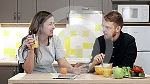 Young couple man and woman with tablet eating breakfast sitting by table in kitchen at home.