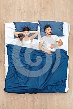 Bedtime. Young man sleeping yawning while woman covering ears angry under blanket on bed top view
