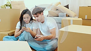 Young couple move into new apartment. Camera moves out of box. Man and woman sit on floor and use smartphones. Loking