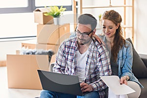 Young couple moving in new home. Sitting and relaxing after unpacking. Searching for ideas on laptop.