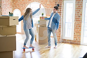 Young couple moving to a new home, having fun riding a skateboard at new apartment around cardboard boxes