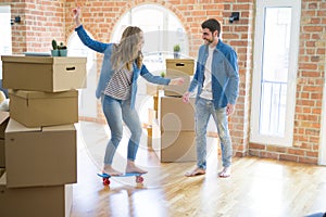 Young couple moving to a new home, having fun riding a skateboard at new apartment around cardboard boxes