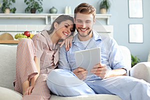 Young couple in pajamas watching media content online in a tablet sitting on a sofa in the living room