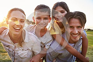 Young couple piggybacking their kids outdoors look to camera