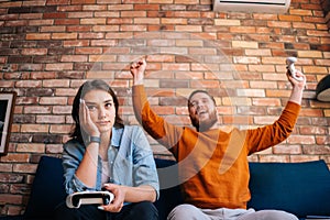 Young couple playing video games on console sitting together on couch at cozy living room. Lady happy to win victory