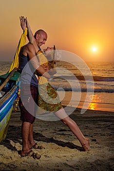 Young couple posing against sea at sunset. Guy and girl fooling and grimace near the old wooden boat on the ocean coast