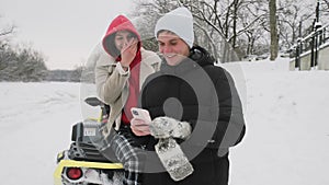 Young couple posing with ATV Quad bike in winter forest. Young man and woman looks at the phone
