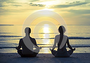 Young couple practicing yoga in the lotus position on the ocean beach during sunset.