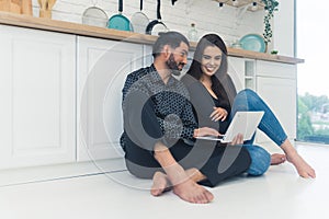 Young couple relaxing barefoot on the floor by kitchen counter using a laptop smiling. Indoor shot.