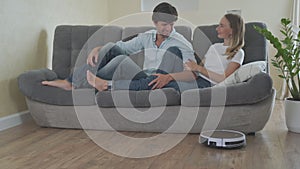 Young couple relaxing on the couch while automatic vacuum cleaner doing the housework in the living room. Household