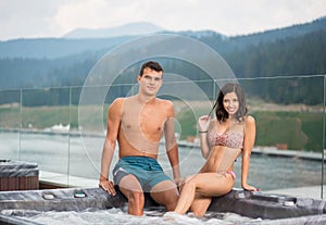 Young couple relaxing enjoying jacuzzi hot tub bubble bath outdoors against background of river, forest and hills
