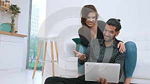 Young couple relaxing in living room together looking at laptop. White woman smiling sitting on couch leaning on her