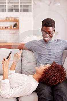 Young Couple Relaxing On Sofa At Home Looking At Mobile Phone Together