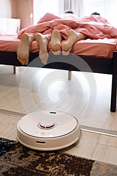 Young couple resting in bed while a robot vacuum cleaner cleans the wooden floor