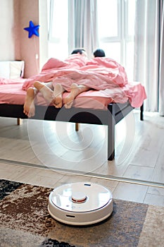 Young couple resting in bed while a robot vacuum cleaner cleans the wooden floor