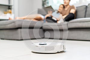 Young couple resting on the couch while robotic vacuum cleaner doing its work at home. Household robots, Smart home concept