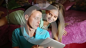 Young couple shopping online using digital tablet computer. Couple laughing, girl lying on the bed at home and guy