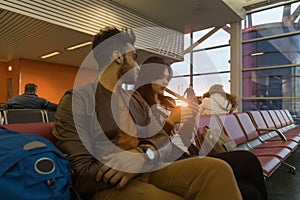 Young Couple Sit In Airport Lounge Waiting Departure Hispanic Man And Woman Use Cell Smart Phone