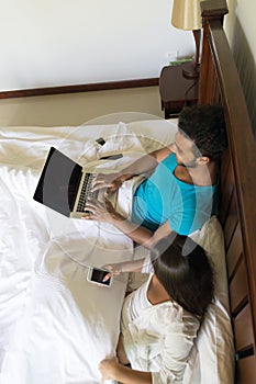 Young Couple Sit In Bed, Happy Smile Hispanic Man And Woman Using Cell Smart Phone Laptop