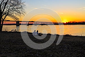 A young couple sitting on the banks of the Mississippi river at sunset near a bridge with cars and trucks driving across