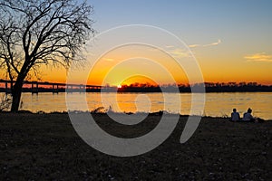 A young couple sitting on the banks of the Mississippi river at sunset near a bridge with cars and trucks driving across