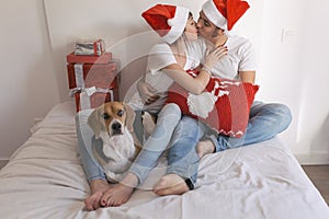 young couple sitting on bed with christmas santa hats having fun with their dog. Presents on bed. Home, indoors. Xmas concept