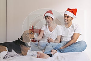 young couple sitting on bed with christmas santa hats having fun with their dog. Presents on bed. Home, indoors. Xmas concept