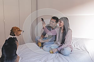 young couple sitting on bed, using mobile phone and having fun. cute beagle dog besides. Breakfast time. Home, indoors