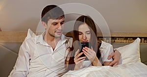Young couple is sitting in bed with white linens. the woman looks intently at her smartphone. the guy comments. they