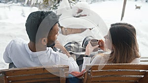 Young couple sitting on the bench outdoors drinking hot chocolate and taking selfie on snowy valentines day