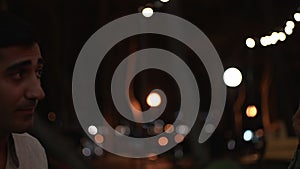 A young couple is sitting in a cafe late at night on blurred garland and black sky background. Media. Close up of man