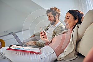 Young couple sitting casually at sofa, smiling, working. Woman looking at her cell phone laughing