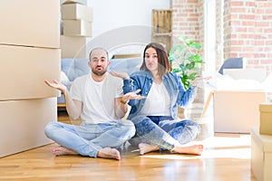 Young couple sitting on the floor arround cardboard boxes moving to a new house clueless and confused expression with arms and