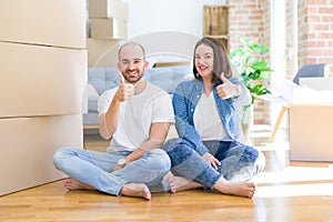 Young couple sitting on the floor arround cardboard boxes moving to a new house doing happy thumbs up gesture with hand