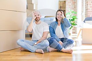 Young couple sitting on the floor arround cardboard boxes moving to a new house doing ok gesture with hand smiling, eye looking