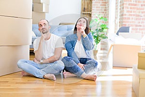 Young couple sitting on the floor arround cardboard boxes moving to a new house with hand on chin thinking about question, pensive